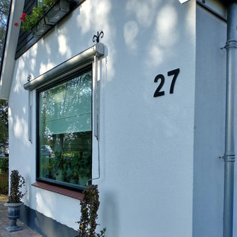 Une maison blanche avec une grande fenêtre, des volets noirs et des lettres adhésives yessential.fr Animo Black 15cm (sans perçage) en gras chiffre 27 sur le mur extérieur. Une petite plante en pot se trouve sous la fenêtre, près du coin.