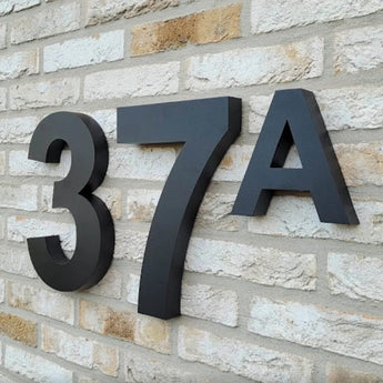 Numéro de maison Angelo Black 30 cm et lettre 37A de yessential.fr dans un style moderne, montés sur un mur de briques de couleur claire.