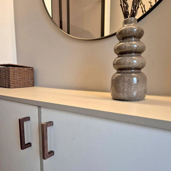 Le buffet Vasco Walnut de yessential.fr, avec poignées en noyer, se dresse sous un miroir mural rond. Un vase texturé marron avec des branches séchées décore le dessus, tandis qu'un panier en osier se trouve à gauche.