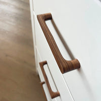 Gros plan de deux poignées en noyer Salgado de yessential.fr, avec une finition lisse, montées sur des tiroirs d'armoire blancs avec un plancher en bois clair en arrière-plan.