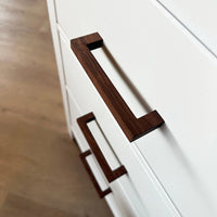 Gros plan de deux tiroirs blancs avec poignées en noyer Vasco de yessential.fr, sur un parquet en bois clair. Ces poignées angulaires modernes sont fabriquées en noyer riche pour une touche élégante.