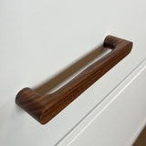 Un gros plan d'une poignée en noyer Salgado de yessential.fr fixée à une armoire blanche, avec un design lisse et moderne et un riche grain de bois brun.