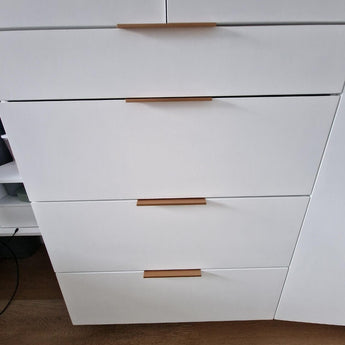 Un gros plan de quatre tiroirs de placard blancs avec des poignées de meuble modernes yessential.fr Enzo Rose Gold dans une finition horizontale, à côté d'une porte de placard assortie partiellement ouverte, le tout sur un parquet en bois.