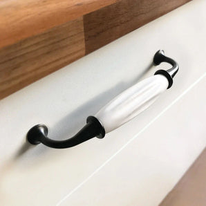 Gros plan de la poignée Daro Ceramic Black de yessential.fr, avec un embout en métal noir et une poignée en céramique blanche, montée sur une façade de tiroir blanche sous un plan de travail en bois.