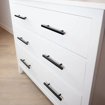 Le buffet Marano Black de yessential.fr dispose de trois tiroirs blancs avec des poignées noires minimalistes, sur un parquet en bois clair contre un mur blanc.