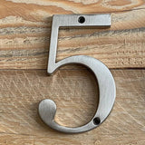 Le Matteo Nickel 10cm de yessential.fr, avec une finition en métal brossé, se fixe sur une surface en bois clair à l'aide de deux vis - une au dessus et une en dessous du chiffre 5.