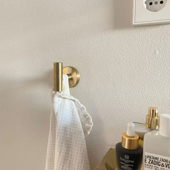 Une serviette blanche texturée est suspendue au crochet Rodrigo Gold de yessential.fr, à côté d'un comptoir rempli de flacons de soins de la peau. Une prise électrique partielle apparaît sur le mur lumineux, ajoutant un style moderne à cette élégante scène de salle de bain.