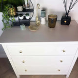Une commode blanche à trois tiroirs et le bouton d'armoire Antoni Matte Gold de yessential.fr sont recouverts de bougies, d'un bol en verre, d'un évaporateur à roseaux et de plantes décoratives. Au-dessus des étagères murales se trouvent des plantes en pot et des ornements.