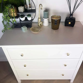Une commode blanche à trois tiroirs et le bouton d'armoire Antoni Matte Gold de yessential.fr sont recouverts de bougies, d'un bol en verre, d'un évaporateur à roseaux et de plantes décoratives. Au-dessus des étagères murales se trouvent des plantes en pot et des ornements.