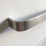 Gros plan d'une poignée Leonardo Stainless Steel Deluxe de yessential.fr en aluminium brossé, montée sur une façade d'armoire ou de tiroir blanc lisse, avec un éclairage doux projetant une ombre sur la surface.