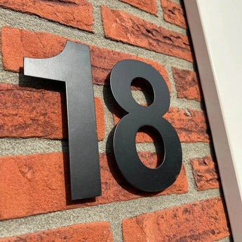 Les numéros de maison Adamo Black 15 cm de yessential.fr sont montés sur un mur de briques rouges avec une bordure blanche, offrant un look de numéro de maison noir moderne et élégant pour votre maison.