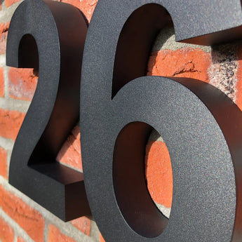 Les numéros de maison Angelo Black 20 cm de yessential.fr ornent le mur de briques rouges avec leur design moderne et leur aspect 3D noir saisissant ; le choix idéal pour un numéro de maison contemporain.