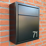 Une boîte aux lettres noire moderne avec le numéro 71, réalisée avec des numéros adhésifs Animo en acier inoxydable de 7,6 cm (sans perçage) de yessential.fr, est montée sur un mur de briques rouges.