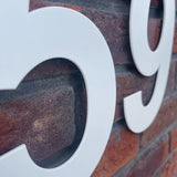 Gros plan des numéros de maison Adamo White de 15 cm de yessential.fr « 5 » et « 9 » sur un mur de briques rouges, avec le numéro 5 au point et une partie du numéro 9 visible à droite.
