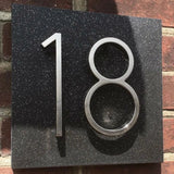 L'Amato Chrome 12,5 cm de yessential.fr est un numéro de maison en métal de style chromé, monté sur une plaque mouchetée noire sur un mur de briques. La lumière du soleil projette des ombres qui accentuent l’aspect tridimensionnel.