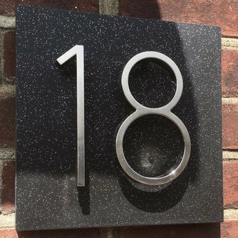 L'Amato Chrome 12,5 cm de yessential.fr est un numéro de maison en métal de style chromé, monté sur une plaque mouchetée noire sur un mur de briques. La lumière du soleil projette des ombres qui accentuent l’aspect tridimensionnel.