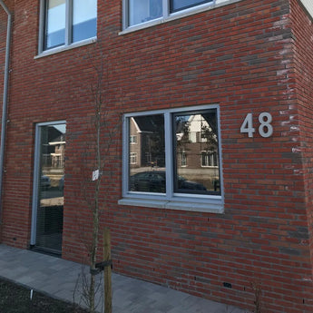 Un bâtiment en brique avec un numéro de maison yessential.fr Enrico en béton de 30 cm au mur, avec deux grandes fenêtres et un petit arbre sur le trottoir pavé. Les reflets des maisons voisines apparaissent dans les fenêtres.