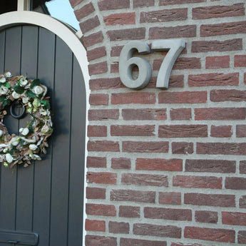 Un gros plan d'un mur de briques montre la maison Enrico en béton de 20 cm numéro 67 de yessential.fr en grands chiffres métalliques, montée à côté d'une porte cintrée gris foncé décorée d'une couronne florale verte et blanche.