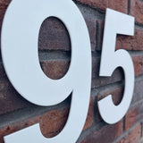 Les grands numéros de maison blancs "95" dans le style Adamo White 20cm de yessential.fr sont montés sur un mur de briques rouges. La photo en angle met en valeur à la fois la texture de la brique et la finition lisse de ce numéro de maison blanc.
