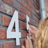 Quelqu'un installe un numéro de maison yessential.fr Adamo White de 20 cm à côté d'un 4 blanc sur un mur de briques rouges. Une partie d'un tuyau métallique apparaît à droite.