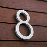 La maison numéro huit Milano de 15 cm en acier inoxydable de yessential.fr avec finition brossée est montée sur un mur en bois avec des lattes horizontales.