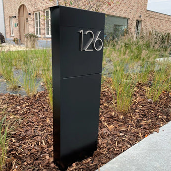 Un numéro de maison Amato Chrome de 12,5 cm de yessential.fr avec le numéro 126 se trouve dans un lit de jardin paillé, entouré d'herbes ornementales devant une maison en brique.