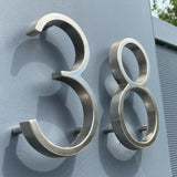 Les numéros Amato Chrome 12,5 cm de yessential.fr sont présentés montés sur un mur gris, photographiés sous un angle pour révéler leur forme tridimensionnelle et leurs ombres spectaculaires.