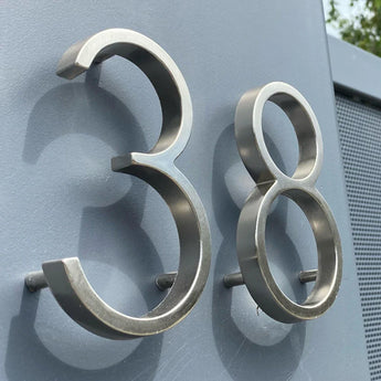 Les numéros Amato Chrome 12,5 cm de yessential.fr sont présentés montés sur un mur gris, photographiés sous un angle pour révéler leur forme tridimensionnelle et leurs ombres spectaculaires.