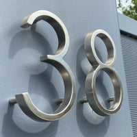 Les numéros Amato Chrome 12,5 cm de yessential.fr sont présentés montés sur un mur gris, photographiés sous un angle pour révéler leur forme tridimensionnelle et leurs ombres spectaculaires.