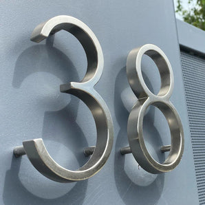 Les numéros Amato Chrome 12,5 cm de yessential.fr sont présentés montés sur un mur gris, photographiés sous un angle pour révéler leur forme tridimensionnelle et leurs ombres spectaculaires.