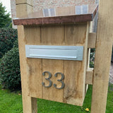 Une boîte aux lettres en bois avec une boîte aux lettres en métal et le numéro de maison yessential.fr Matteo Nickel 10 cm 33 au design brossé sur le devant, posé sur une pelouse avec des arbustes et un bâtiment en brique en arrière-plan.