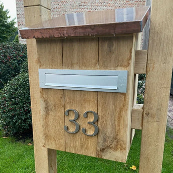 Une boîte aux lettres en bois avec une boîte aux lettres en métal et le numéro de maison yessential.fr Matteo Nickel 10 cm 33 au design brossé sur le devant, posé sur une pelouse avec des arbustes et un bâtiment en brique en arrière-plan.