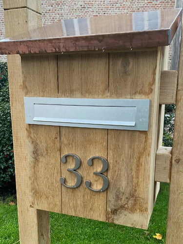 Une boîte aux lettres en bois avec une boîte aux lettres en métal et le numéro de maison yessential.fr Matteo Nickel 10 cm 33 au design brossé sur le devant, posé sur une pelouse avec des arbustes et un bâtiment en brique en arrière-plan.
