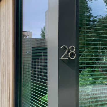 Une façade moderne avec une grande fenêtre présente le numéro de maison Amato Chrome de 12,5 cm de yessential.fr, monté sur un panneau vertical sombre à côté du verre. Les stores et la verdure sont visibles par la fenêtre.