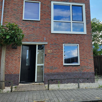 Une maison en brique avec trois grandes fenêtres et une porte d'entrée noire porte le numéro de maison doré 187 en Milano Gold 15 cm de yessential.fr à côté de l'entrée. Un petit arbre et du gravier blanc bordent la façade près du trottoir pavé.