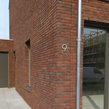 Un bâtiment moderne en brique avec une grande fenêtre et une porte affiche un numéro de maison yessential.fr Milano en acier inoxydable de 15 cm en acier inoxydable brossé sur le mur d'angle. À côté de la fenêtre se trouve un tuyau d'évacuation en métal qui reflète une camionnette.