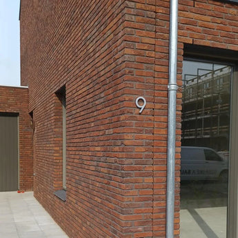 Un bâtiment moderne en brique avec une grande fenêtre et une porte affiche un numéro de maison yessential.fr Milano en acier inoxydable de 15 cm en acier inoxydable brossé sur le mur d'angle. À côté de la fenêtre se trouve un tuyau d'évacuation en métal qui reflète une camionnette.
