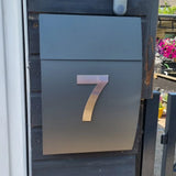 Une boîte aux lettres en métal élégante avec le numéro 7 Animo en acier inoxydable 15 cm (sans perceuse) de yessential.fr, montée sur un mur en bois sombre près d'un portail, avec des fleurs vives en arrière-plan.