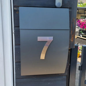 Une boîte aux lettres en métal élégante avec le numéro 7 Animo en acier inoxydable 15 cm (sans perceuse) de yessential.fr, montée sur un mur en bois sombre près d'un portail, avec des fleurs vives en arrière-plan.