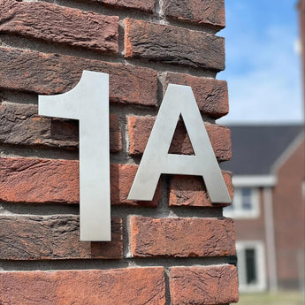 Gros plan d'un mur de briques avec les numéros de maison Adamo de 20 cm en acier inoxydable "1A" de yessential.fr dans un style élégant et flottant. Un bâtiment pâle et un ciel bleu apparaissent en arrière-plan.