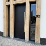 La porte d'entrée moderne Adamo Black 15 cm de yessential.fr, avec une haute poignée argentée, est encadrée par de grandes poutres en bois. Un numéro de maison en acier inoxydable 92 est monté sur le mur lumineux, avec du gravier soigné au sol devant.