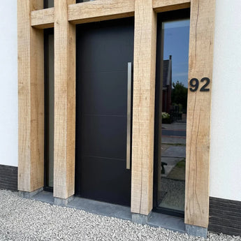 La porte d'entrée moderne Adamo Black 15 cm de yessential.fr, avec une haute poignée argentée, est encadrée par de grandes poutres en bois. Un numéro de maison en acier inoxydable 92 est monté sur le mur lumineux, avec du gravier soigné au sol devant.
