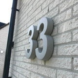 Les grands numéros de maison Enrico gris en béton de 30 cm de yessential.fr sont montés sur un mur de briques claires. La photo, prise sous un angle, souligne l'épaisseur 3D et projette des ombres saisissantes sur la surface.