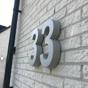 Les grands numéros de maison Enrico gris en béton de 30 cm de yessential.fr sont montés sur un mur de briques claires. La photo, prise sous un angle, souligne l'épaisseur 3D et projette des ombres saisissantes sur la surface.
