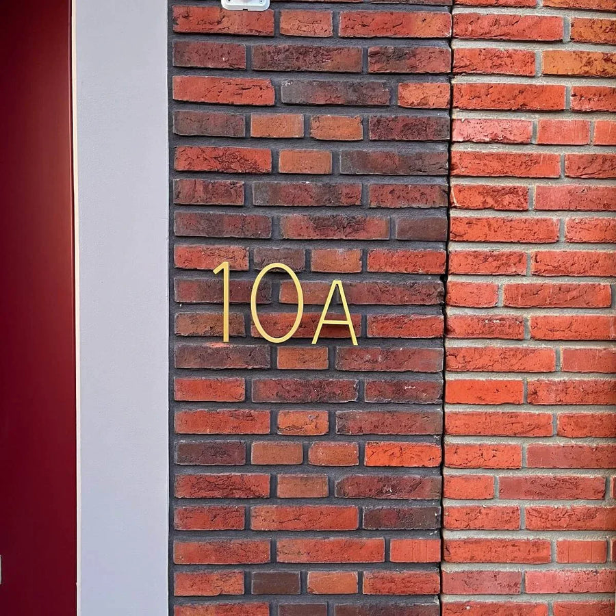 Le numéro et la lettre Amato Gold 15 cm de yessential.fr sont conçus comme des numéros de maison modernes et montés sur un mur de briques marron et rouge à côté d'une bande verticale gris clair et d'une partie de cadre de porte rouge sur la gauche.