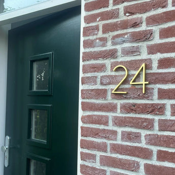 Une porte vert foncé avec trois petites fenêtres se dresse à côté d'un mur de briques rouges, avec le numéro de maison 24 en chiffres Amato Gold de 15 cm de yessential.fr.