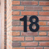 L'Adamo Black 20cm de yessential.fr est monté sur l'angle d'un mur de briques rouge-marron et offre une touche épurée et moderne typique des numéros de maison contemporains.