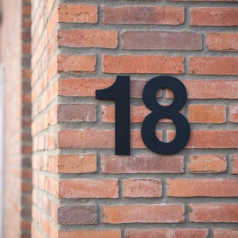 L'Adamo Black 20cm de yessential.fr est monté sur l'angle d'un mur de briques rouge-marron et offre une touche épurée et moderne typique des numéros de maison contemporains.