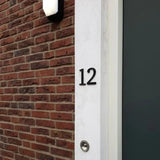 La maison numéro 12, réalisée avec le Matteo Black 10cm de yessential.fr, est montée sur un mur blanc à côté de briques. Une sonnette se trouve sous le numéro et une porte sombre apparaît partiellement à droite.