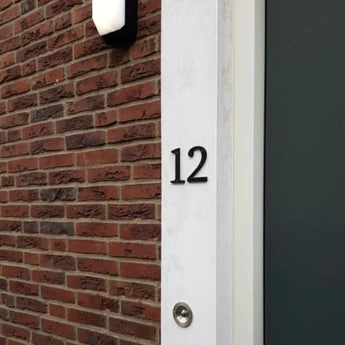 La maison numéro 12, réalisée avec le Matteo Black 10cm de yessential.fr, est montée sur un mur blanc à côté de briques. Une sonnette se trouve sous le numéro et une porte sombre apparaît partiellement à droite.